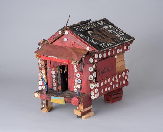 A small sculpture by Beverly Buchanan titled "Shot Gun House." It's mostly painted a dark barn red, made of and decorated with found materials including buttons and a Georgia license plate for the roof. A sign with the words "shot gun" appears on the side.
