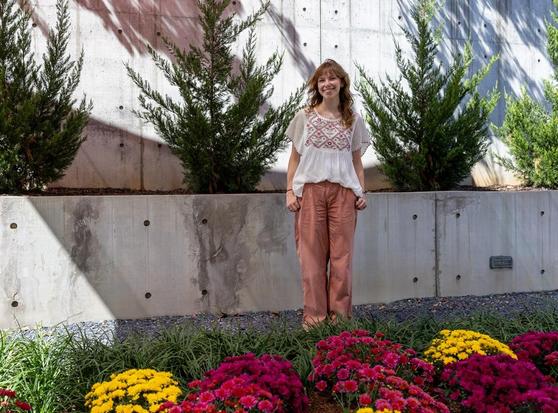 Museum programs assistant Ellen Patton stands for a photo in the museum's Jane and Harry Willson Sculpture Garden, with an array of pink and yellow flowers in front of her and a low concrete wall behind her.