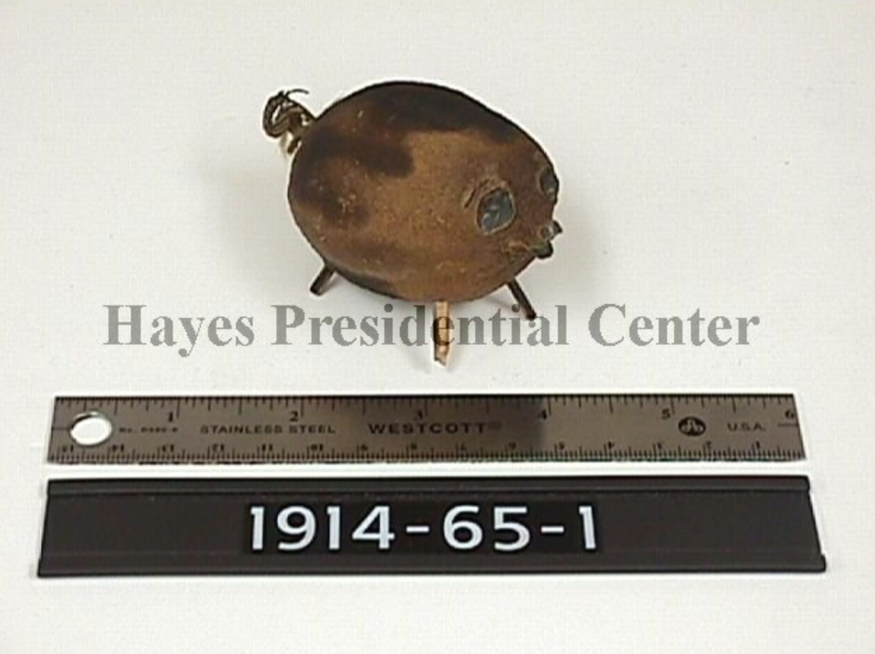 A dried out old lemon that was clearly made into a pig. It has matchstick legs and a tail made out of something. Is is standing next to a ruler and the image has Hayes Presidential Center watermarked across it.