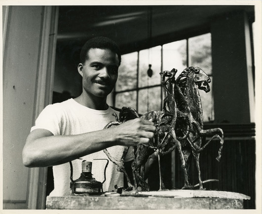 A black-and-white photo of the sculptor John Rhoden, wearing a white t-shirt, and with his hands on his sculpture "Three Horses." It's a tabletop metal sculpture of three horses. A window in the background lets in some light.