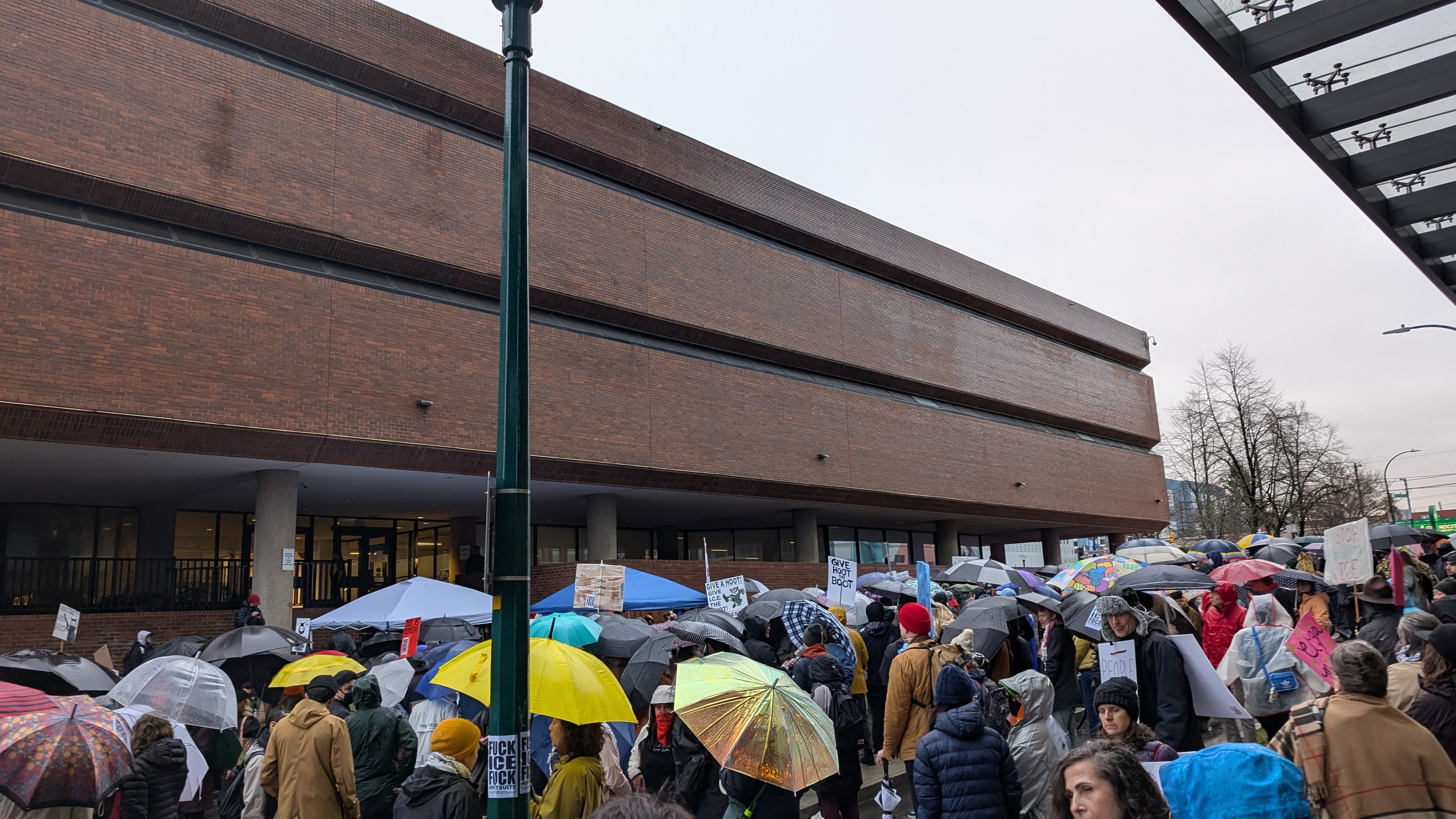 Photo of an outdoor crowd in front of a brutalist brick building. It's a protest with many people holding umbrellas or protest signs