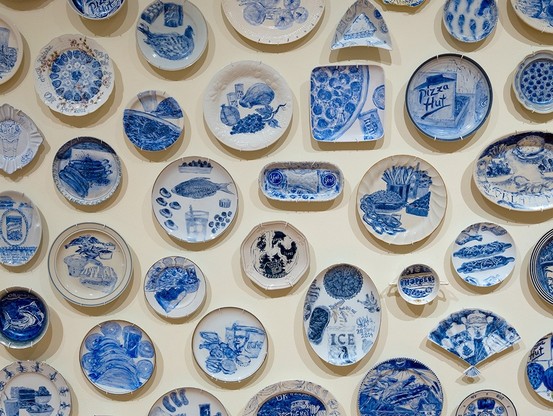 A detail of Julie Green's "The Last Supper," an art project in which Green painted the requested last meals of incarcerated people on death row in blue on found ceramic plates. The image shows about 30 plates, hung close together on a gallery wall.